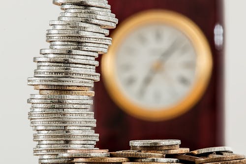 Free A close-up image of stacked coins with a blurred clock, symbolizing time and money relationship. Stock Photo